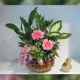 Pink gerbera daisies in a glass bowl with lush green foliage and a small bird figurine