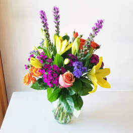 Mixed bouquet of yellow lilies, roses, and purple flowers in a glass vase
