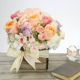 Low wooden box arrangement of pastel roses and hydrangeas with a cream bow on a table.