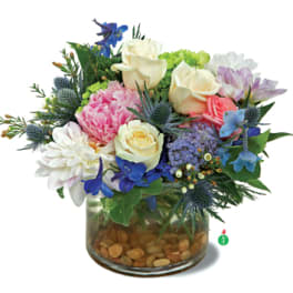 Low glass vase of mixed cream roses, pink peony, white dahlia, and blue and purple flowers over river stones