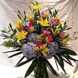 Tall arrangement of yellow lilies, pink roses, and pale lavender hydrangeas in a glass vase
