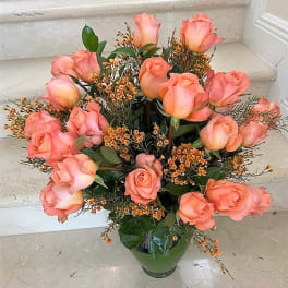 Tall arrangement of peach roses with tiny orange filler blooms in a green glass vase