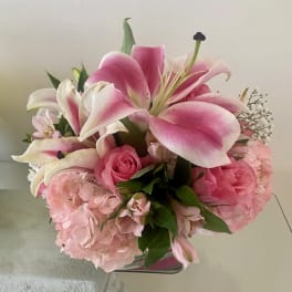 Pink and white lilies, roses, and hydrangeas arranged in a short glass cube vase