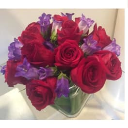 Compact arrangement of red roses and purple flowers in a clear glass cube vase.