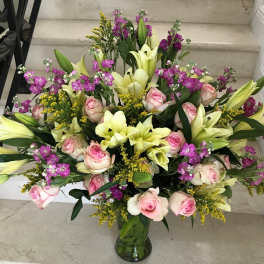 Large mixed bouquet of pale yellow lilies, pink roses, and purple flowers in a clear glass vase