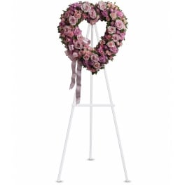 Heart-shaped wreath of pink and lavender roses on a white standing easel with a satin ribbon bow.