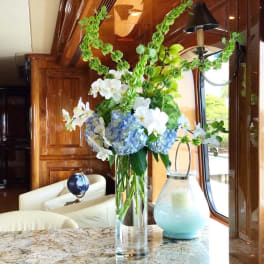 Tall floral arrangement with blue hydrangeas, white orchids, and green hanging garlands in a glass vase