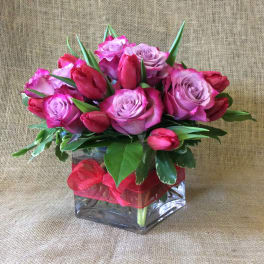 Pink roses and red tulips arranged in a square glass vase with a red ribbon bow.