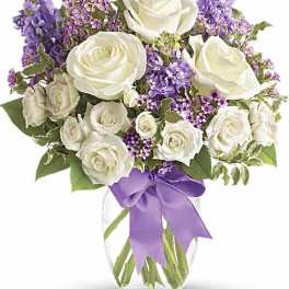 Arrangement of white roses with purple flowers in a clear vase with a lavender bow