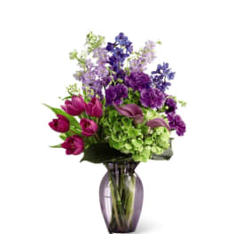 Tall arrangement of purple blooms, green hydrangea, and magenta tulips in a purple glass vase