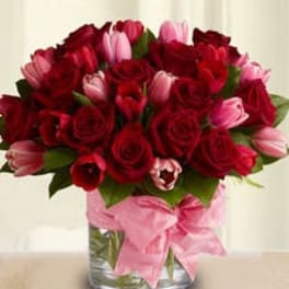 Round arrangement of red roses and pink tulips in a clear glass vase with a pink ribbon bow