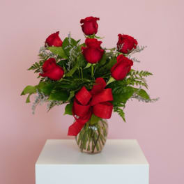 Red rose arrangement in a clear glass vase with a red bow on a white pedestal and pink background