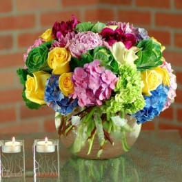 Colorful mixed bouquet of roses, hydrangeas, peonies, and calla lilies in a round glass vase with two votive candles.