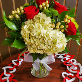 Vase arrangement with white hydrangeas, red roses, green mums, and berries on a table with heart garland.