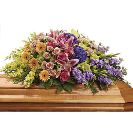 Colorful casket spray of pink lilies, roses, yellow gerbera daisies, and purple flowers on a wooden casket