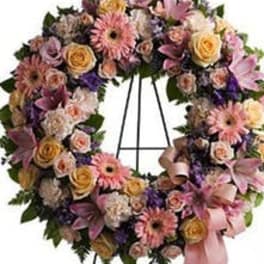 Pastel wreath of pink gerberas, roses, lilies, and carnations on a stand with a pink ribbon bow
