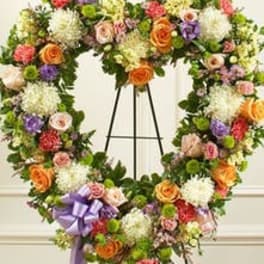 Heart-shaped standing wreath of mixed roses, mums, and other flowers with a lavender bow.