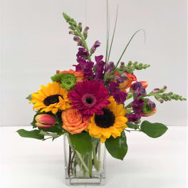 Mixed bouquet in a clear glass vase with sunflowers, roses, and purple spikes