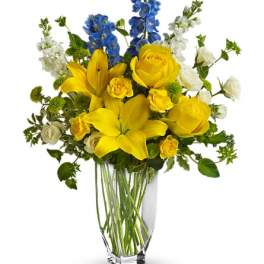 Yellow lilies and roses with blue and white flowers in a clear vase