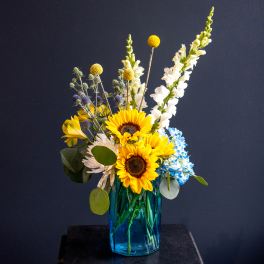 Sunflowers, white snapdragons, and blue hydrangeas in a blue glass vase