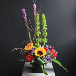 Tall mixed bouquet with sunflowers, red blooms, and green accents in a black vase