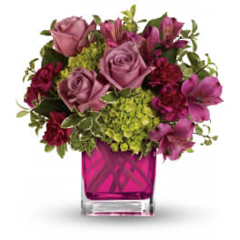 Compact bouquet of purple roses, pink blooms, and green hydrangea in a pink glass cube vase