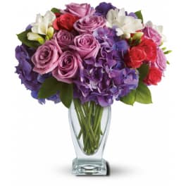 Mixed bouquet of purple hydrangeas, lavender roses, red roses, and white freesias in a clear glass vase.