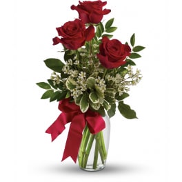 Three red roses with white filler flowers in a clear glass vase with a red ribbon bow