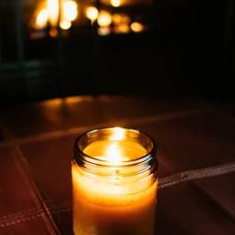 Lit amber candle in a small glass jar on a dark surface with warm light in the background
