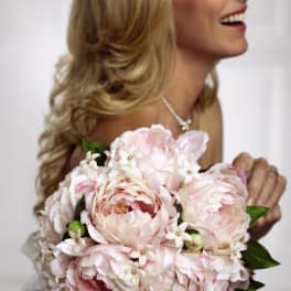 Round bouquet of pale pink peonies with small white blossoms held by a smiling bride.