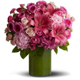 Pink lilies, roses, and peonies arranged in a round bouquet in a green-lined glass cylinder vase