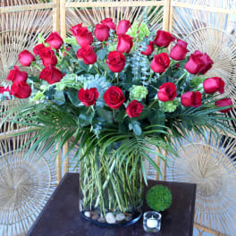 Red roses arranged in a clear glass vase with greenery and palm fronds