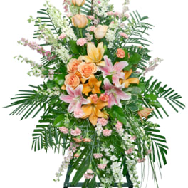Tall standing spray of peach roses, lilies, and pink carnations with cascading white blooms on a green easel.