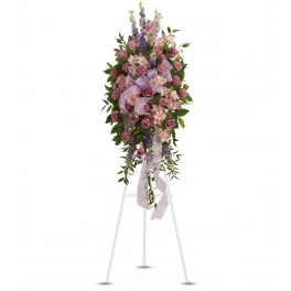 Tall standing spray of mauve roses and pink flowers with purple delphinium and a lavender bow on a white easel.