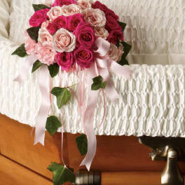 Pink and blush rose bouquet with trailing ribbons and ivy resting on a white casket interior