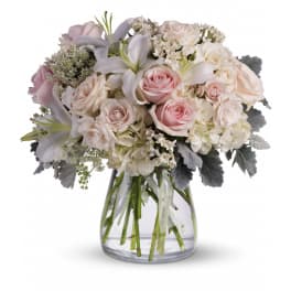 Arrangement of pale pink roses, white lilies, and ivory blooms with soft gray foliage in a clear glass vase