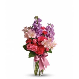 Tall bouquet of pink roses, red carnations, and purple stock in a clear vase with pink bow