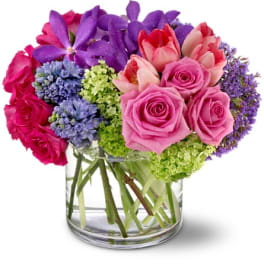 Compact bouquet of pink roses, purple orchids, tulips, and mixed blooms in a clear glass cylinder vase