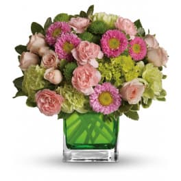 Short arrangement of pink roses and carnations with green blooms in a green glass cube vase