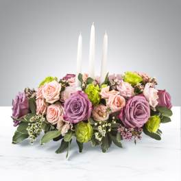 Low centerpiece of pink and purple roses with green blooms surrounding three white taper candles