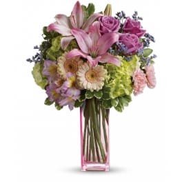 Tall arrangement of pink lilies, lavender roses, and mixed pastel flowers in a pink glass vase