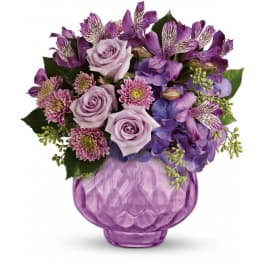 Lavender roses, purple alstroemeria, mums, and hydrangea arranged in a round purple glass vase