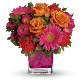 Compact bouquet of orange roses, pink gerbera daisies and red flowers in a bright pink glass cube vase