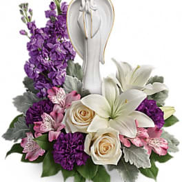 White angel figurine surrounded by white lilies, cream roses, and purple and pink flowers