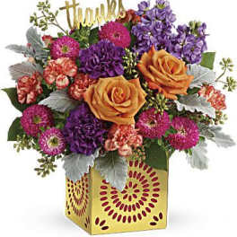 Bright mixed arrangement of orange roses, purple and pink flowers in a yellow cube vase with a gold “thanks” sign