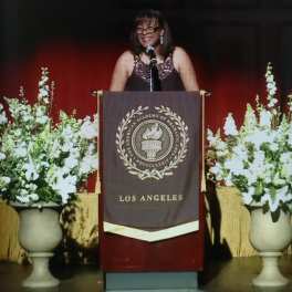 White floral arrangements in urn vases flank a podium on a stage