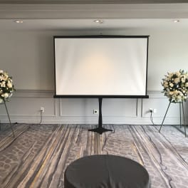 Two white floral standing arrangements flank a projection screen in a room