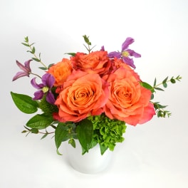 Orange roses in a white vase with purple flowers and greenery