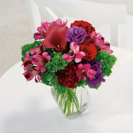 Compact bouquet of red, pink, and purple flowers with bright green accents in a clear glass vase