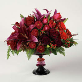 Low arrangement of red roses, dark pink lilies, and red blooms in a black pedestal vase